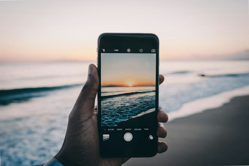 A hand holding up a phone to photograph the sun setting over the ocean